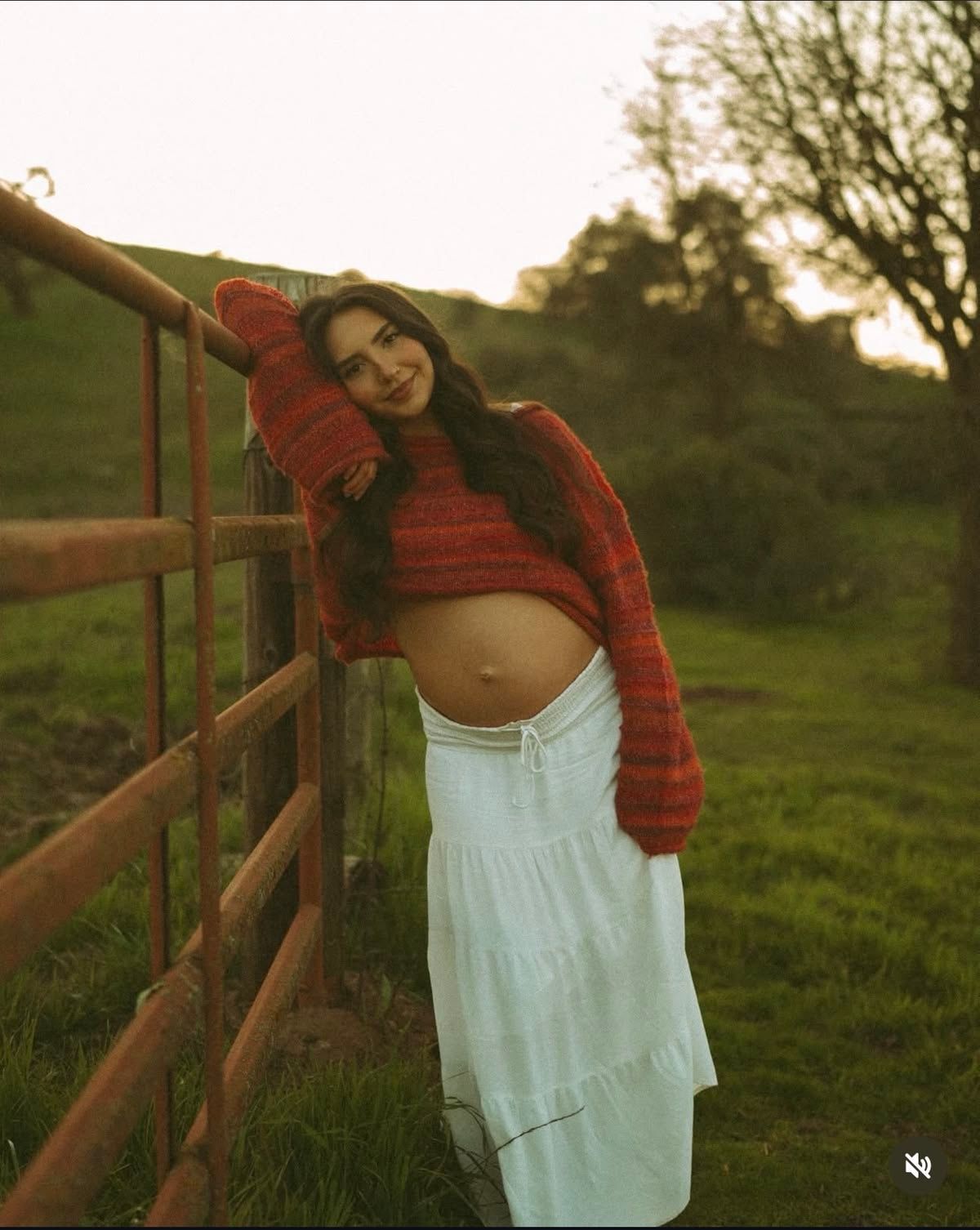 Pregnant woman in red crop sweater and white maxi skirt leaning on wooden fence in green meadow at sunset.