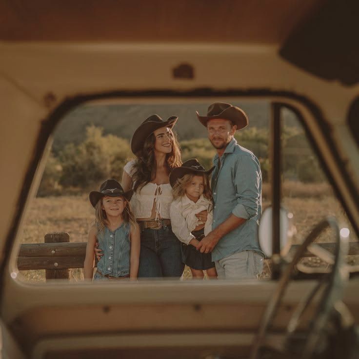 Family of four in cowboy hats posing outdoors, photographed through a vintage vehicle window in a rustic countryside setting.
