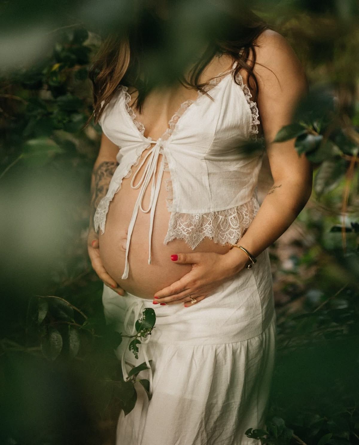 Pregnant woman in white lace dress cradling baby bump outdoors among green leaves, maternity photography.