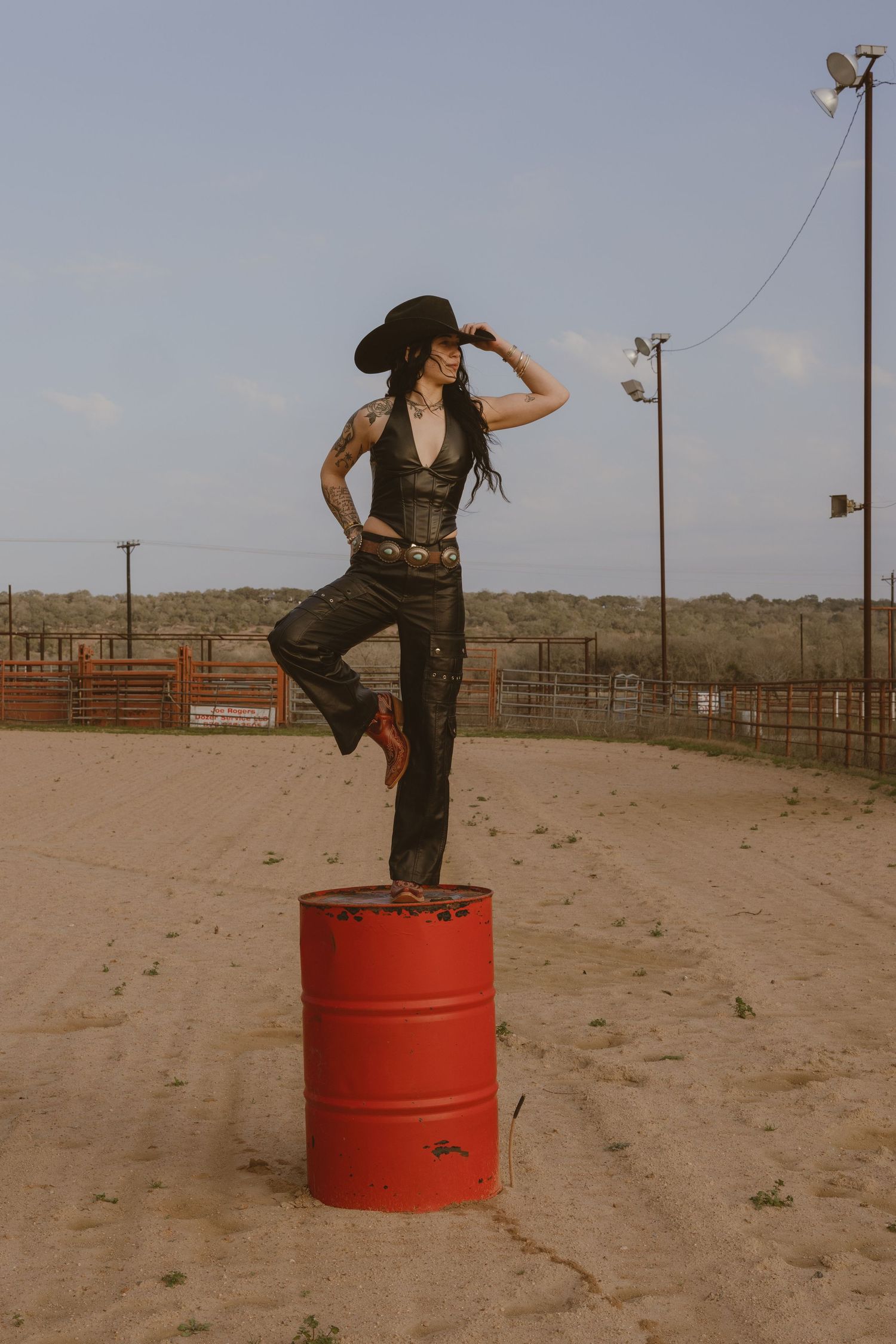 A person in western attire practices mounting a red barrel in a dusty rodeo arena.