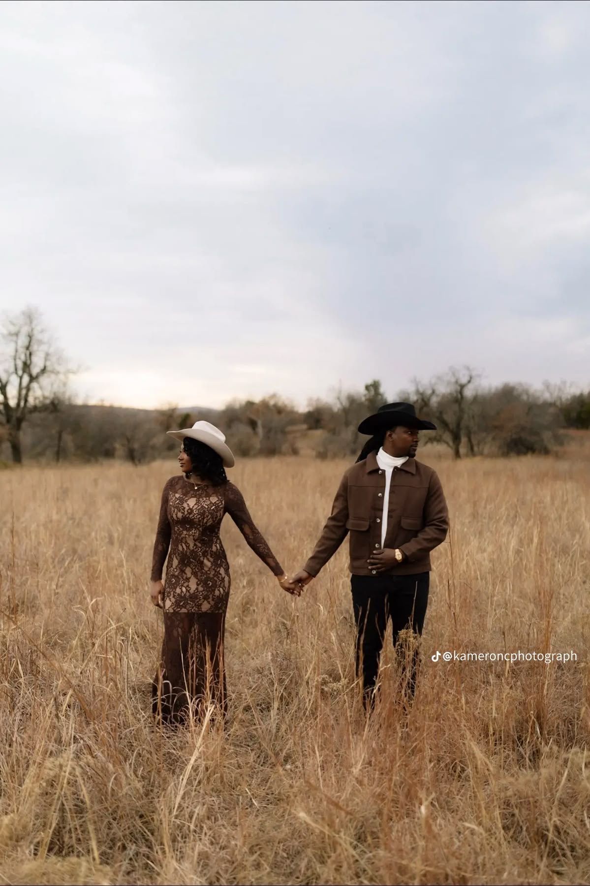 A Black couple in western hats holds hands in a golden field, wearing brown outfits against a cloudy sky.