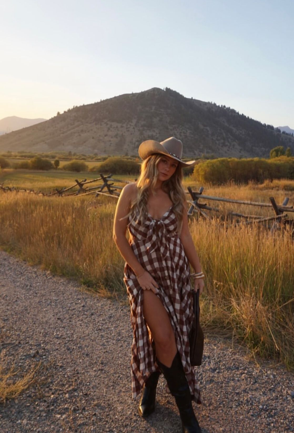 A figure in a plaid dress and wide-brimmed hat poses on a gravel path at sunset with mountains in the background.