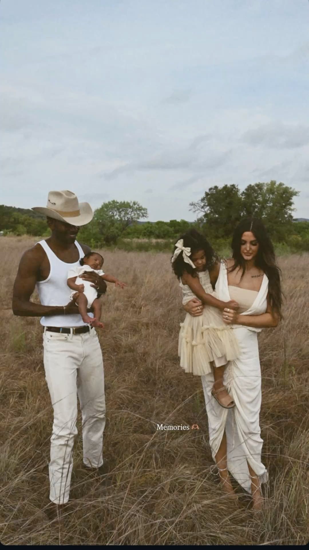 Family of four in white outfits posing in a golden field, with parents holding two young children under a cloudy sky.