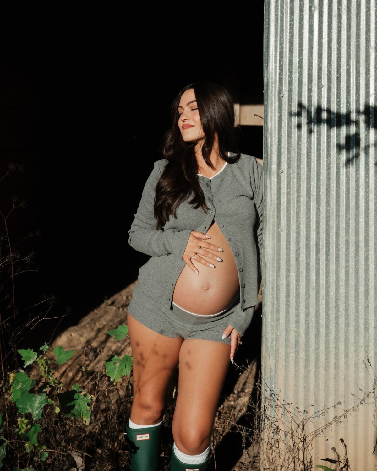 Pregnant woman in grey outfit and green boots leaning against corrugated metal wall outdoors.