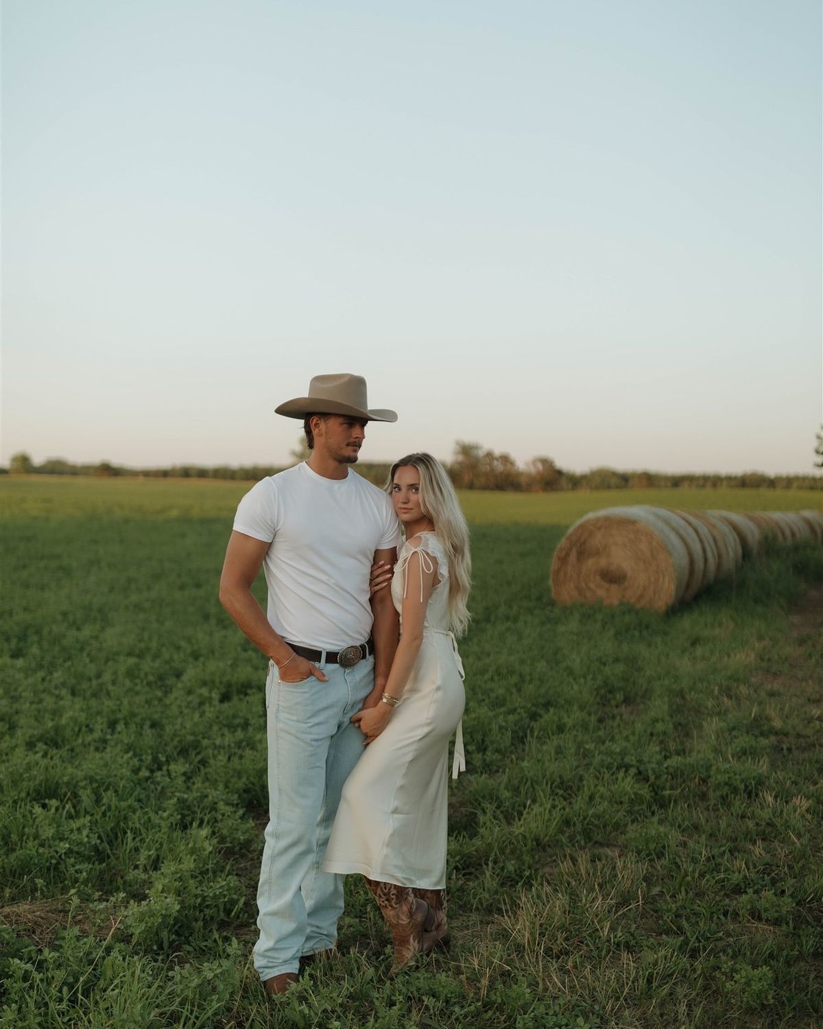 A couple in wedding attire embrace in a rustic field with hay bales at sunset creating a romantic countryside scene.