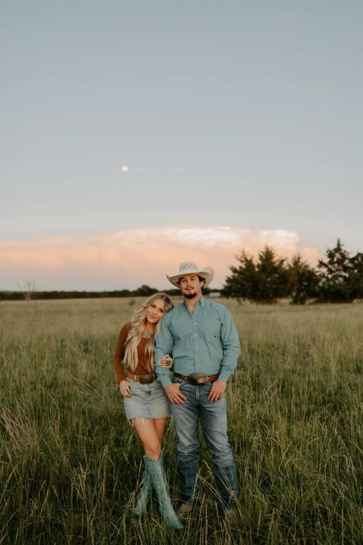 A couple in western attire embraces while standing in a golden prairie field at sunset.