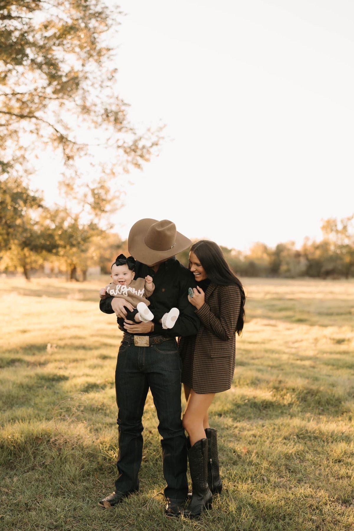 A couple in western attire holds their baby in a sunlit field during golden hour.