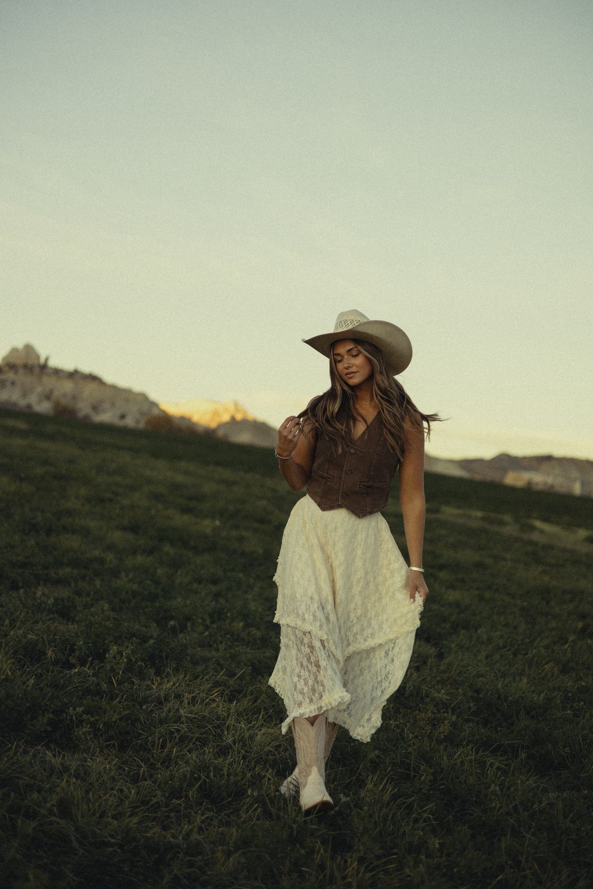 Woman in cowboy hat, brown vest, and white lace skirt walking on a grassy hill at sunset.