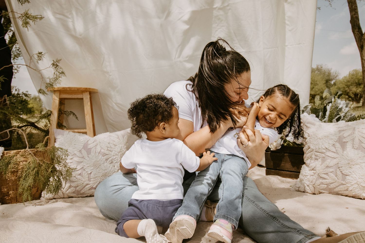A mother joyfully plays with her two young children sitting on cushions in a cozy boho outdoor setting.