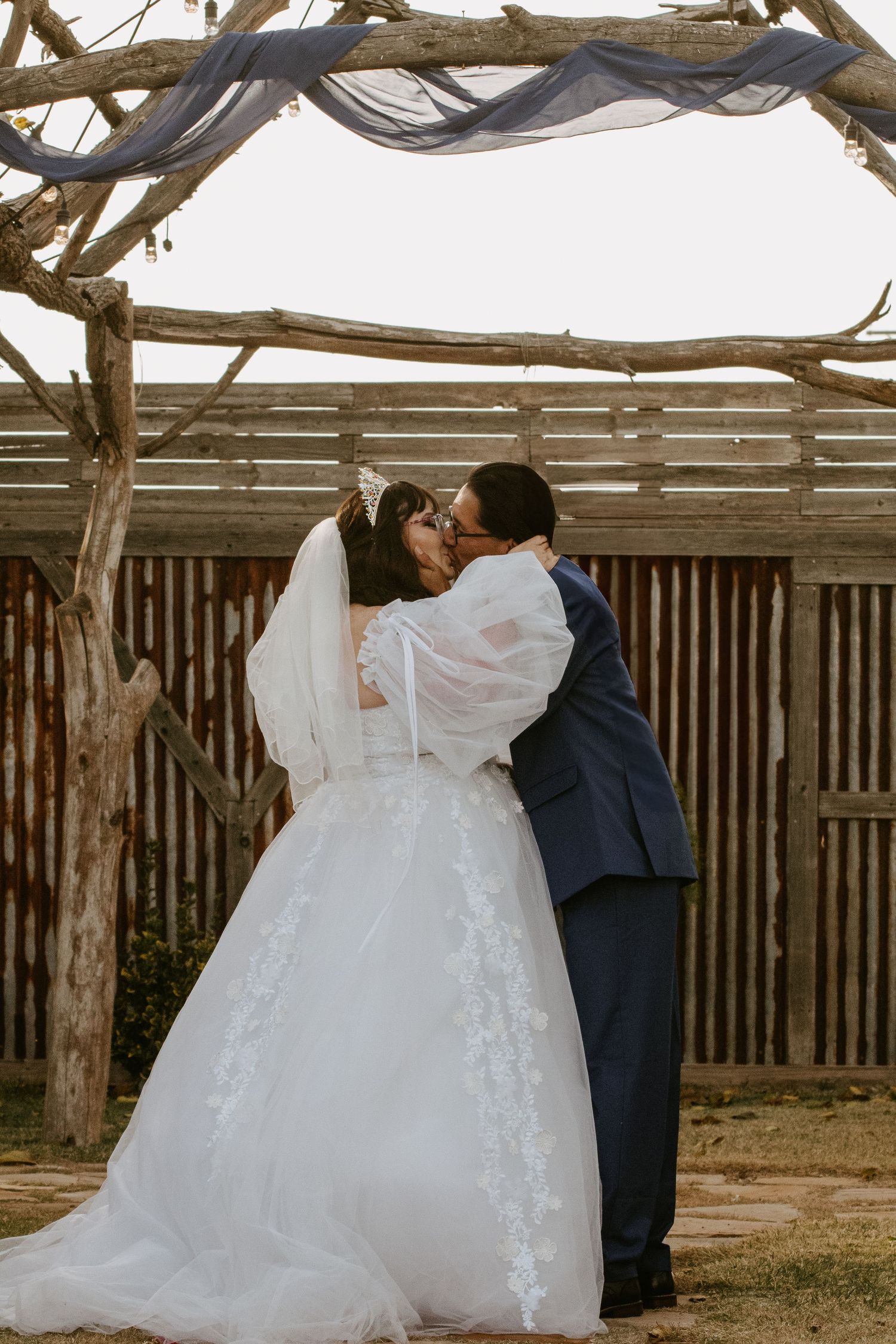 Bride and groom share first kiss under rustic wooden arch adorned with blue fabric at outdoor wedding ceremony.