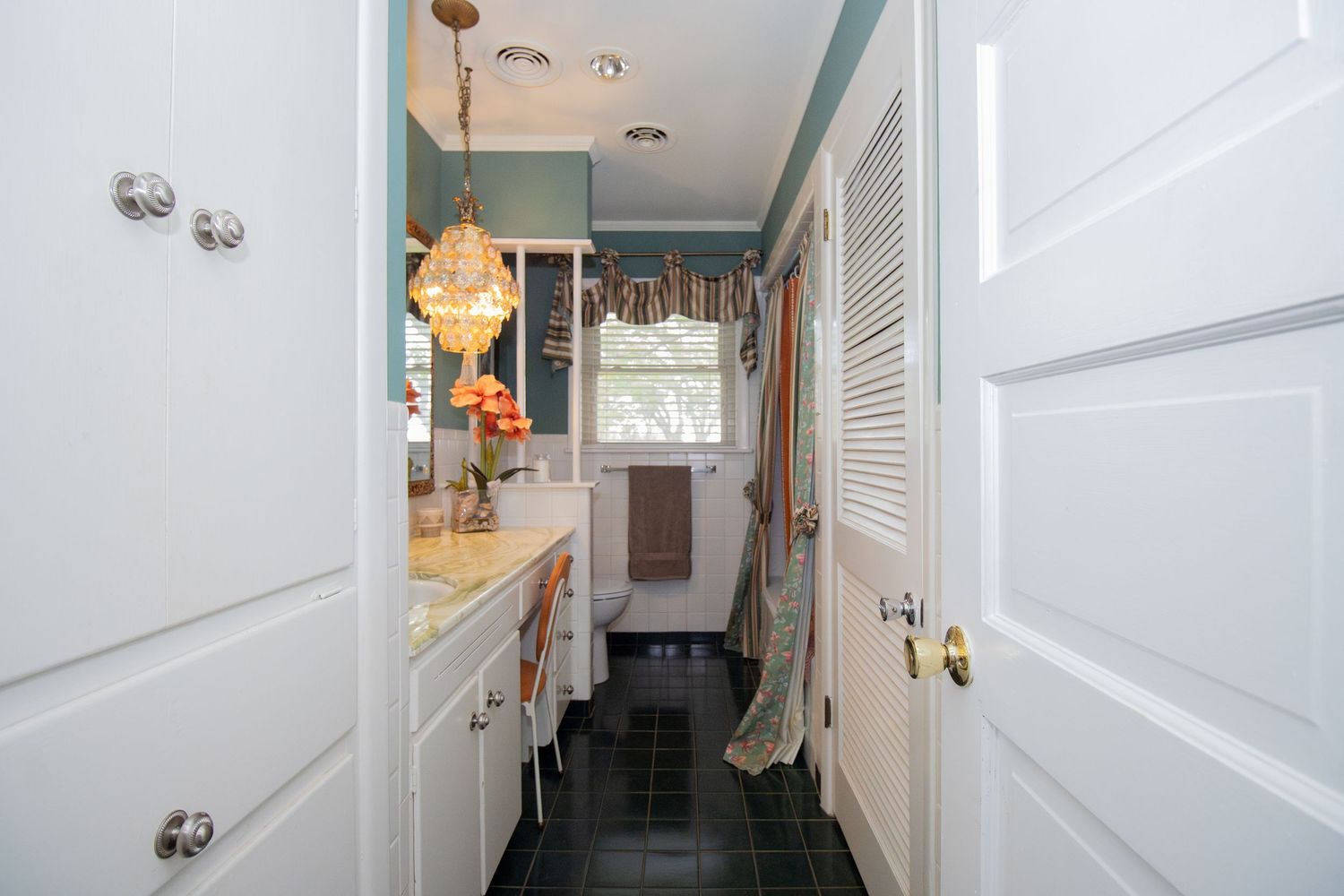 Narrow bathroom with dark tile floors, white cabinetry, teal walls, pendant light, and a window with colorful valance.