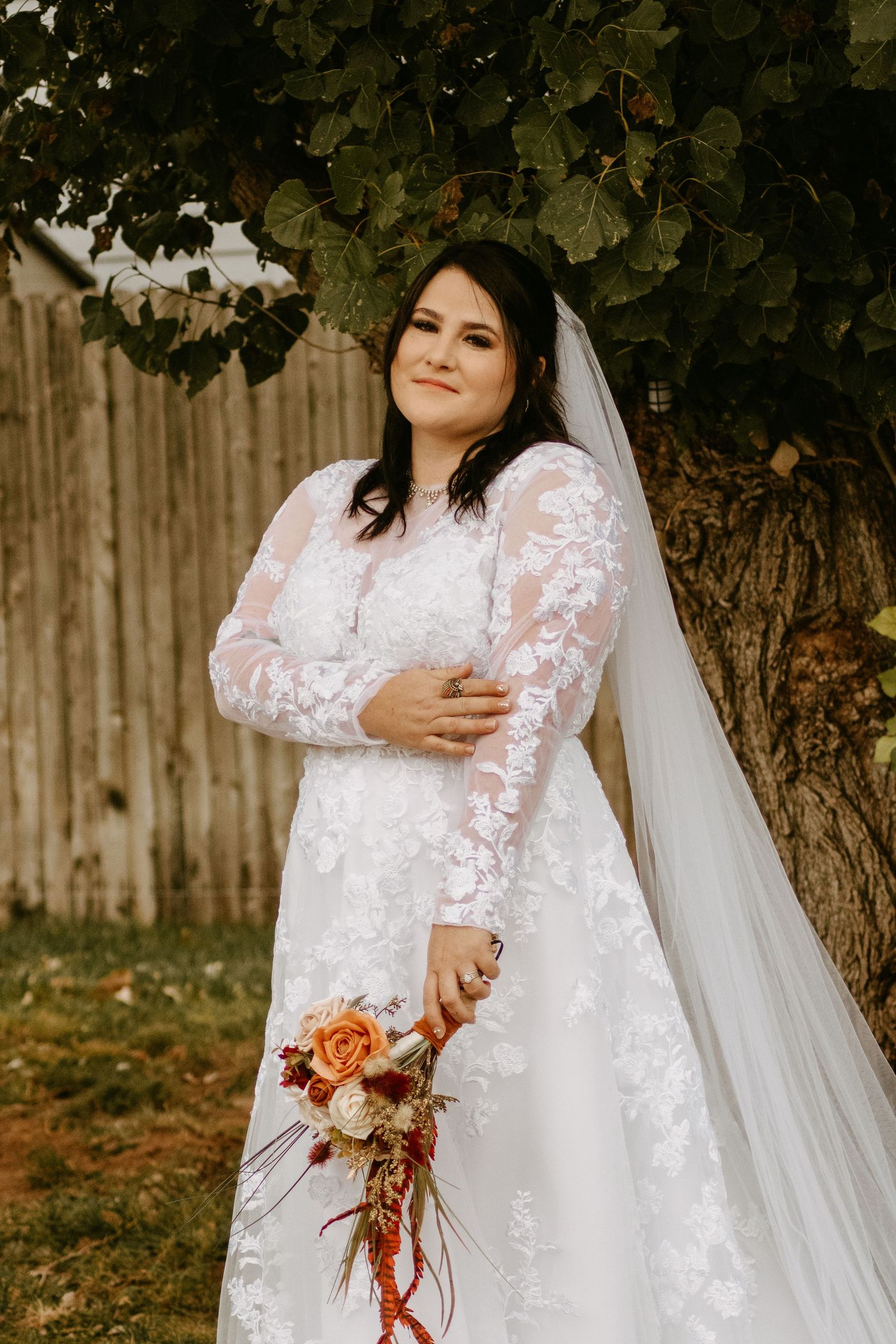 Bride in lace long-sleeve wedding gown holding autumn bouquet, standing under tree with veil flowing, outdoor bridal portrait.