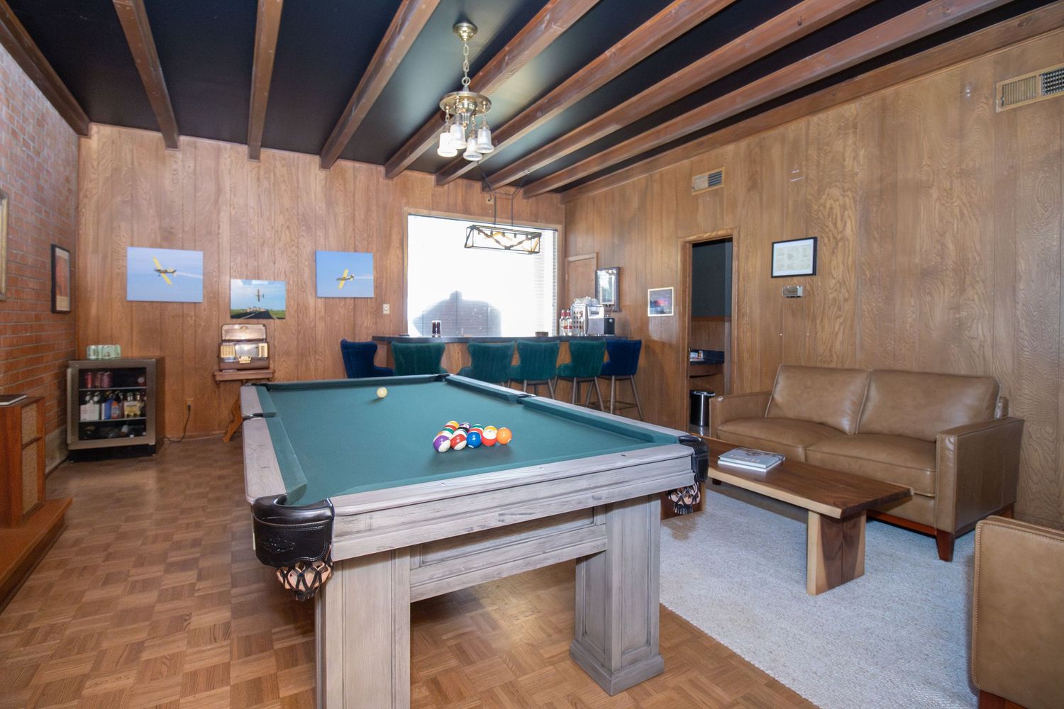 Wood-paneled game room featuring a pool table with billiard balls, leather sofa, chandelier, and hardwood floors.