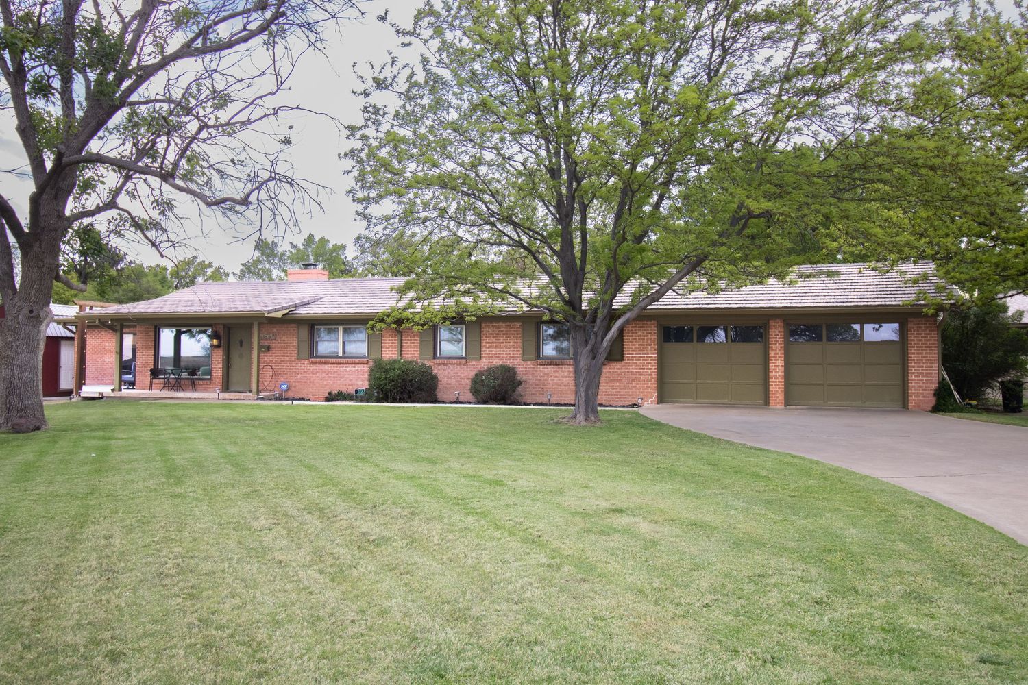 Brick ranch-style home with attached garage, lush green lawn, and large shade trees on a sunny day.