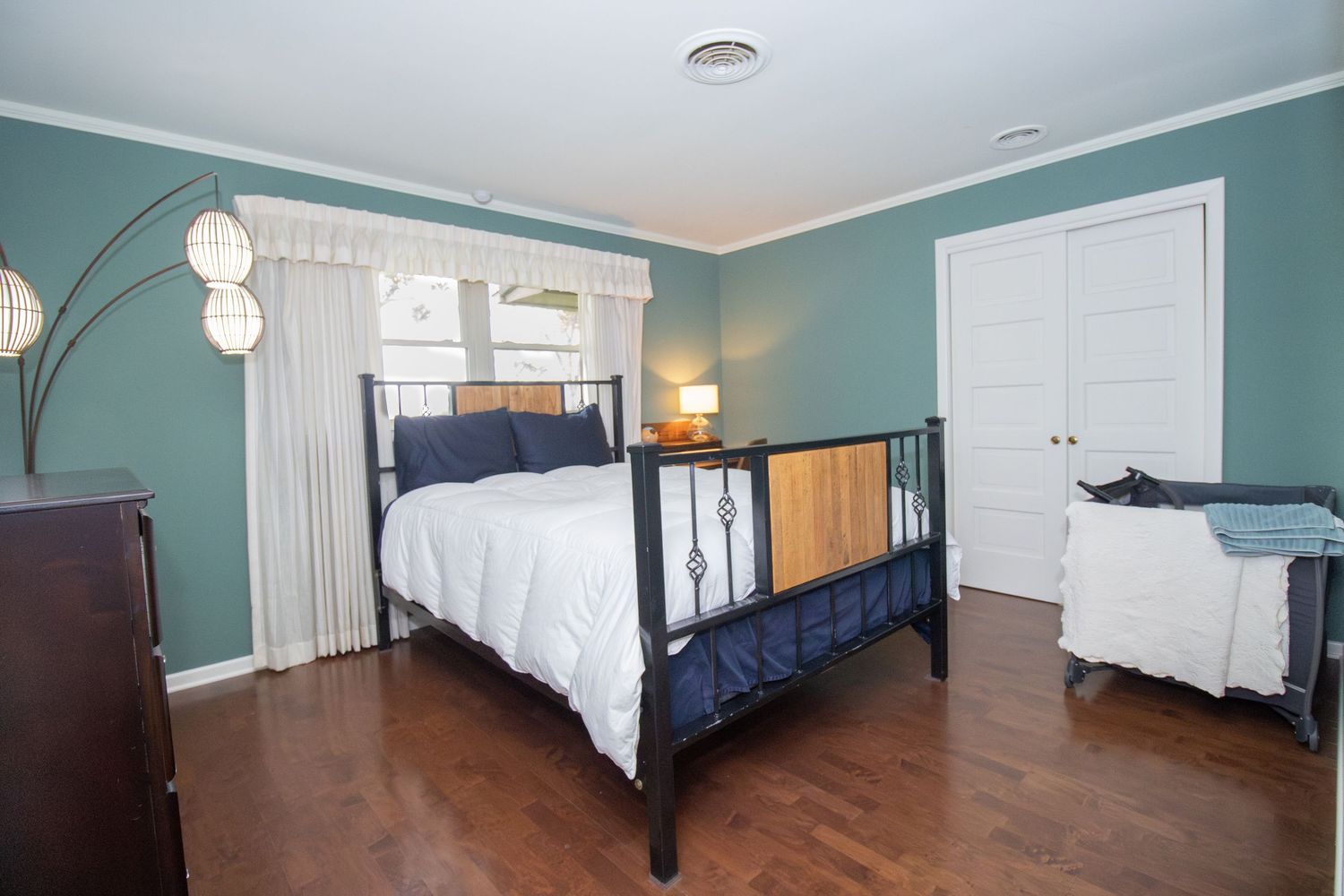 Spacious bedroom with teal walls, dark hardwood floors, black metal bed frame, white bedding, and a modern arc floor lamp.