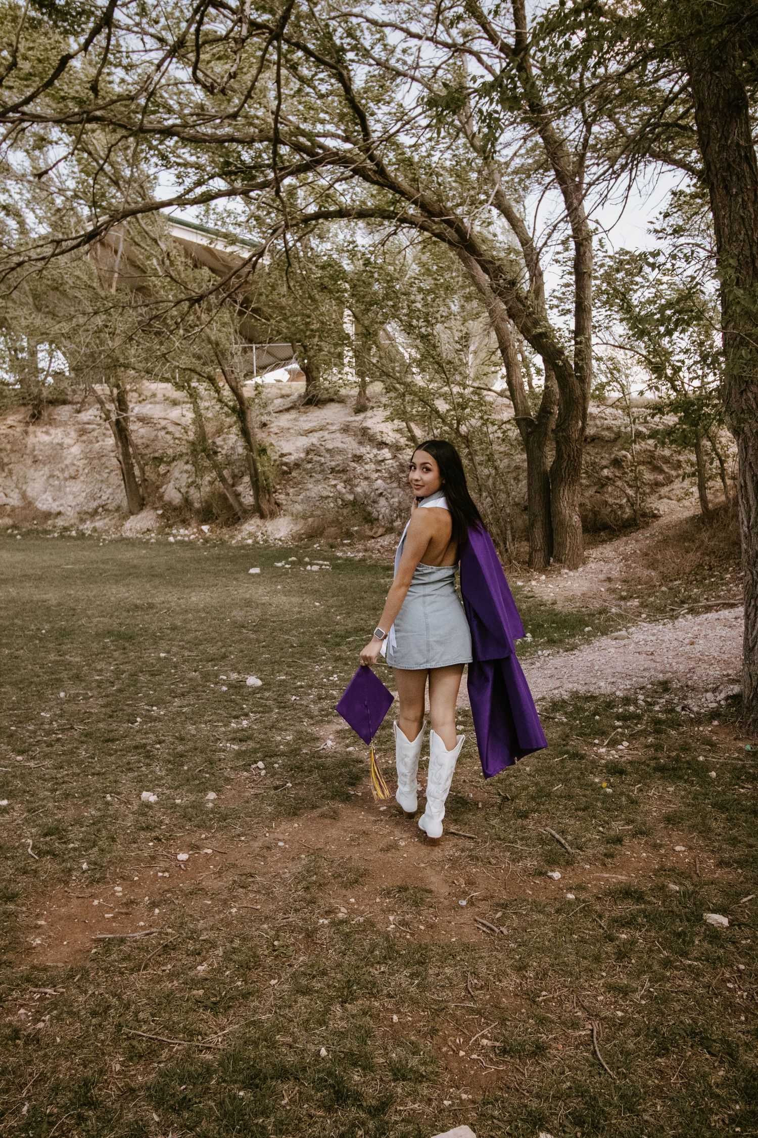 Young woman in white cowboy boots and mini dress wearing purple graduation gown walks outdoors among trees.