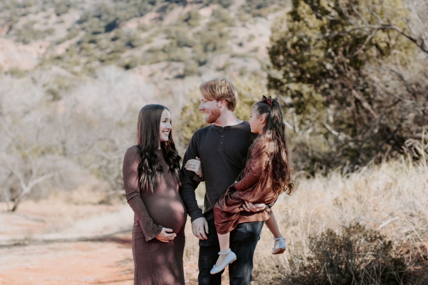 Pregnant woman and man holding child outdoors on a desert trail with dry brush and mountains in background.