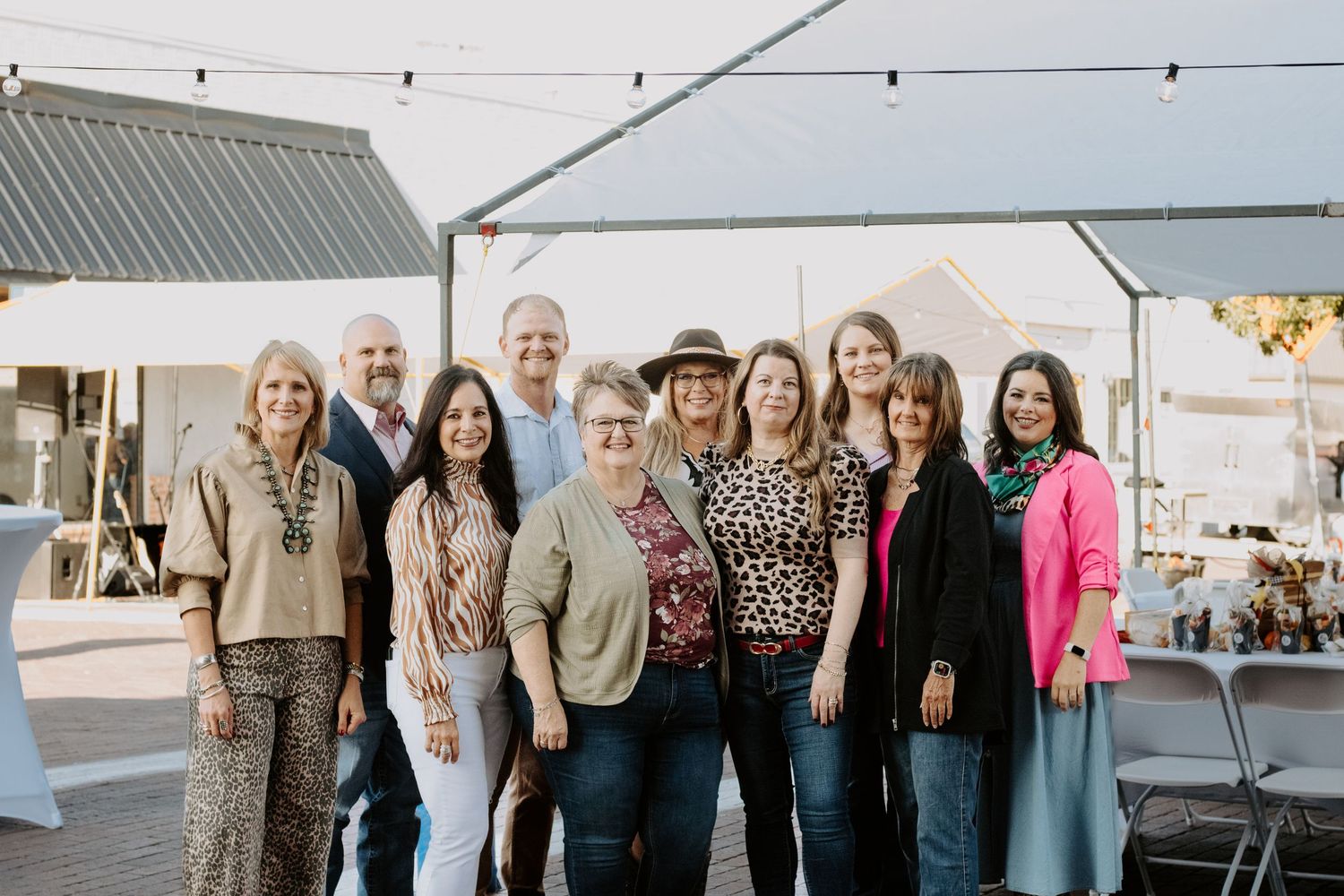 Group of ten smiling adults posing together under a white tent at an outdoor event.
