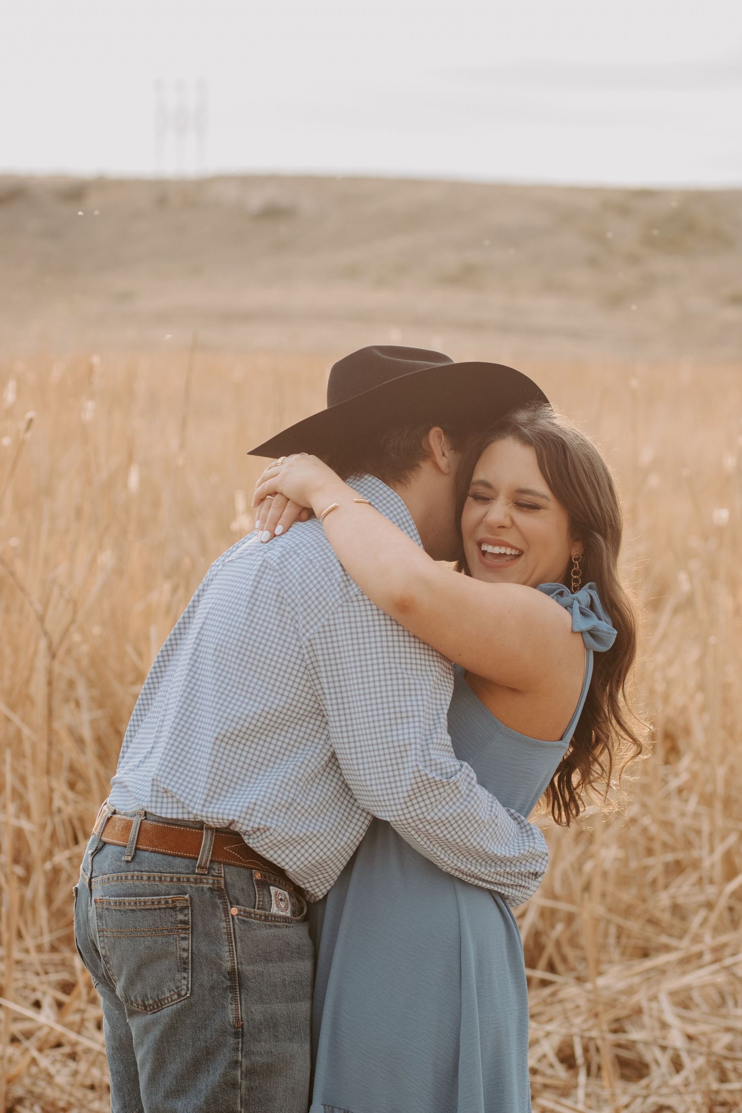 A couple embraces joyfully in a golden field, the man wearing a cowboy hat and the woman in a blue dress.