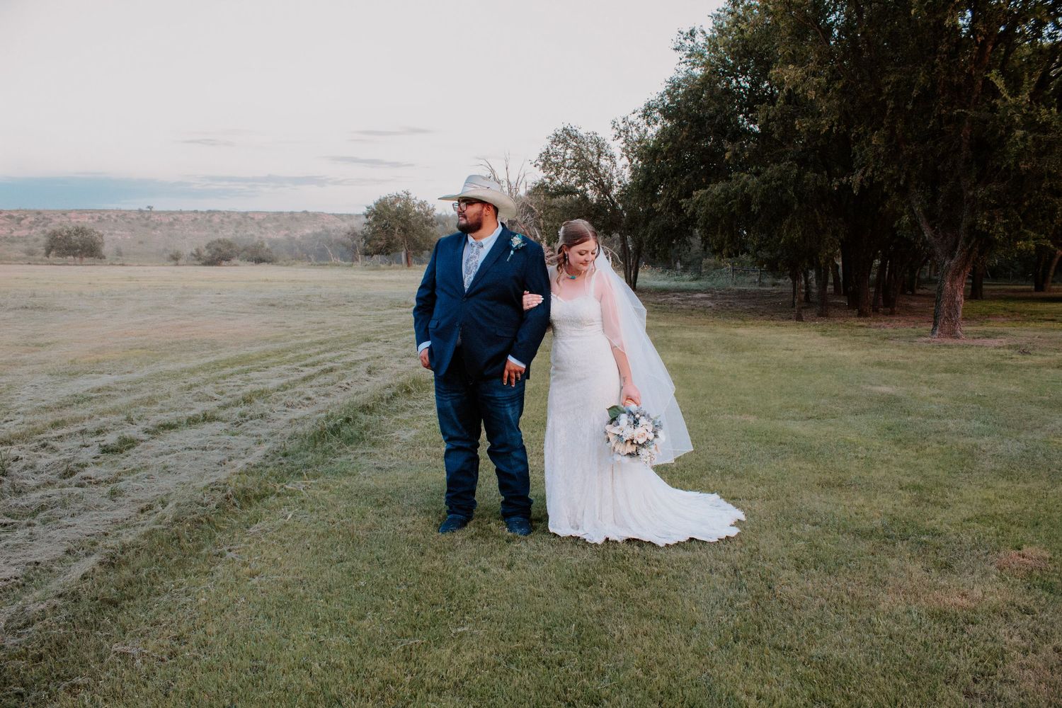 Bride in white gown and groom in blue suit with cowboy hat pose on green field at rustic outdoor wedding.