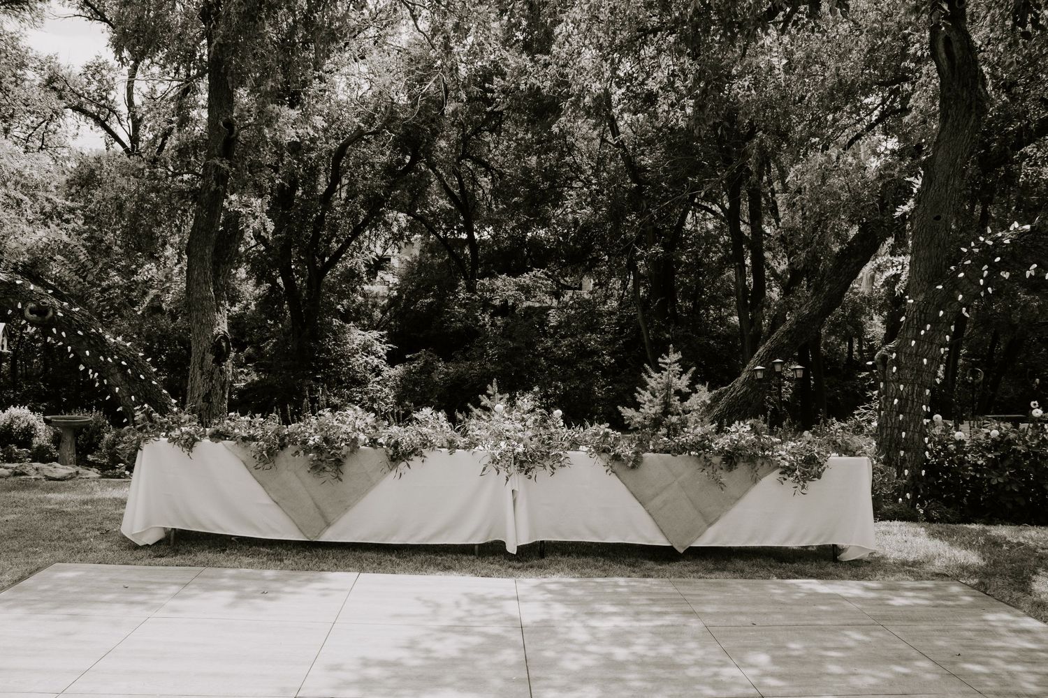 Elegant outdoor wedding head table with white linen and floral garland, surrounded by trees with string lights.