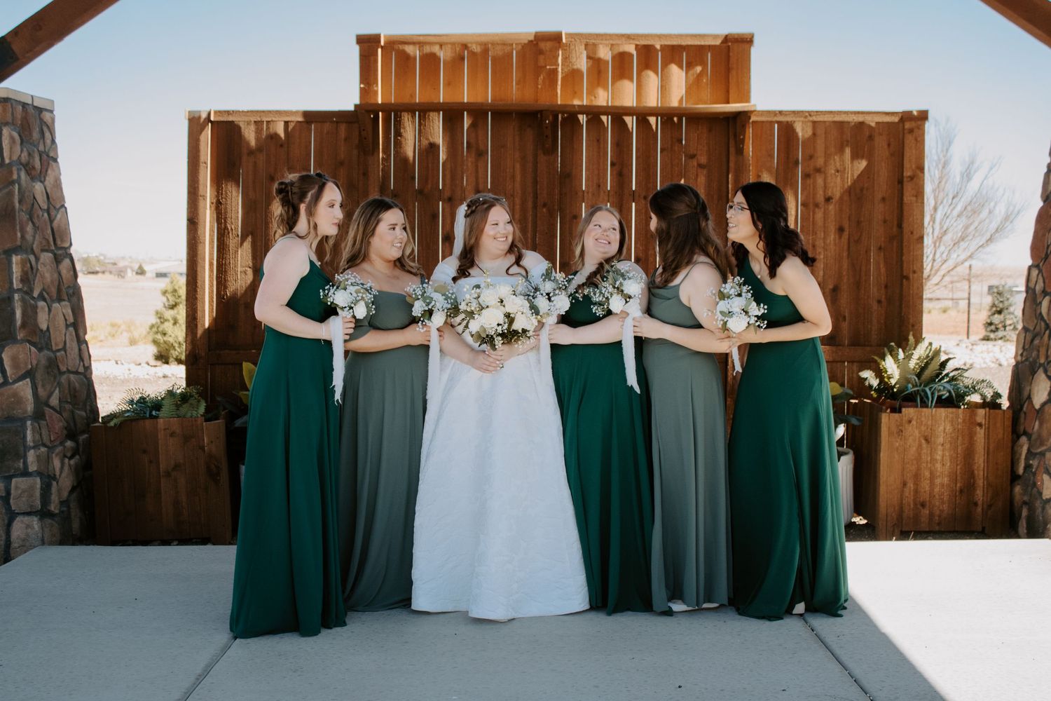 Bride in white gown with bridesmaids in emerald green dresses holding bouquets outside rustic barn wedding venue.