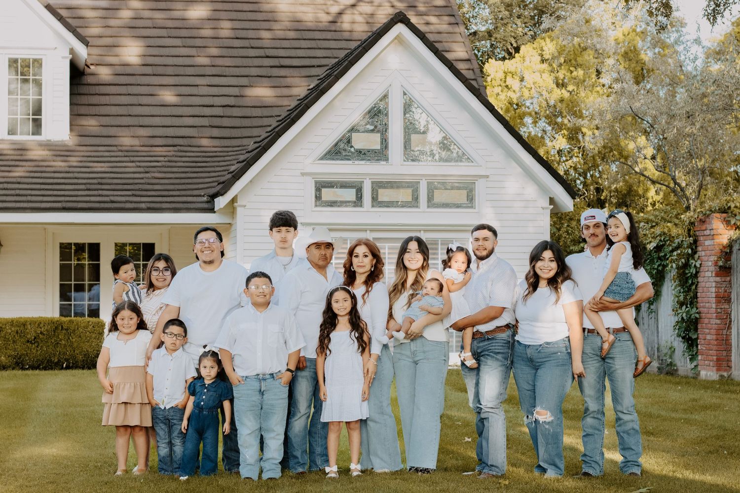 Large multigenerational family posing together outdoors in front of a white farmhouse wearing coordinated white and denim outfits.