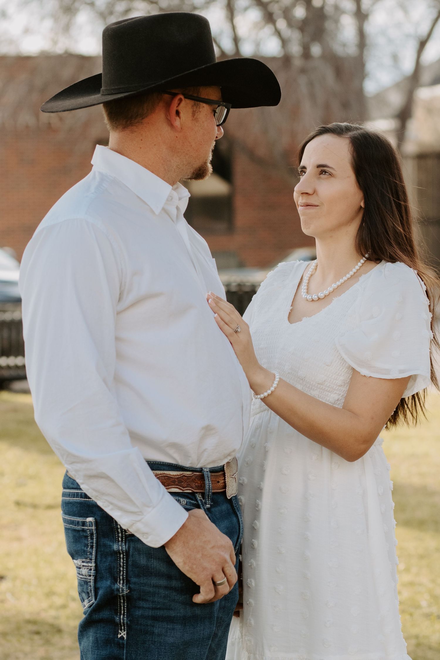 Couple in matching white outfits, man in cowboy hat and jeans, woman in white dress with pearl necklace, outdoors.