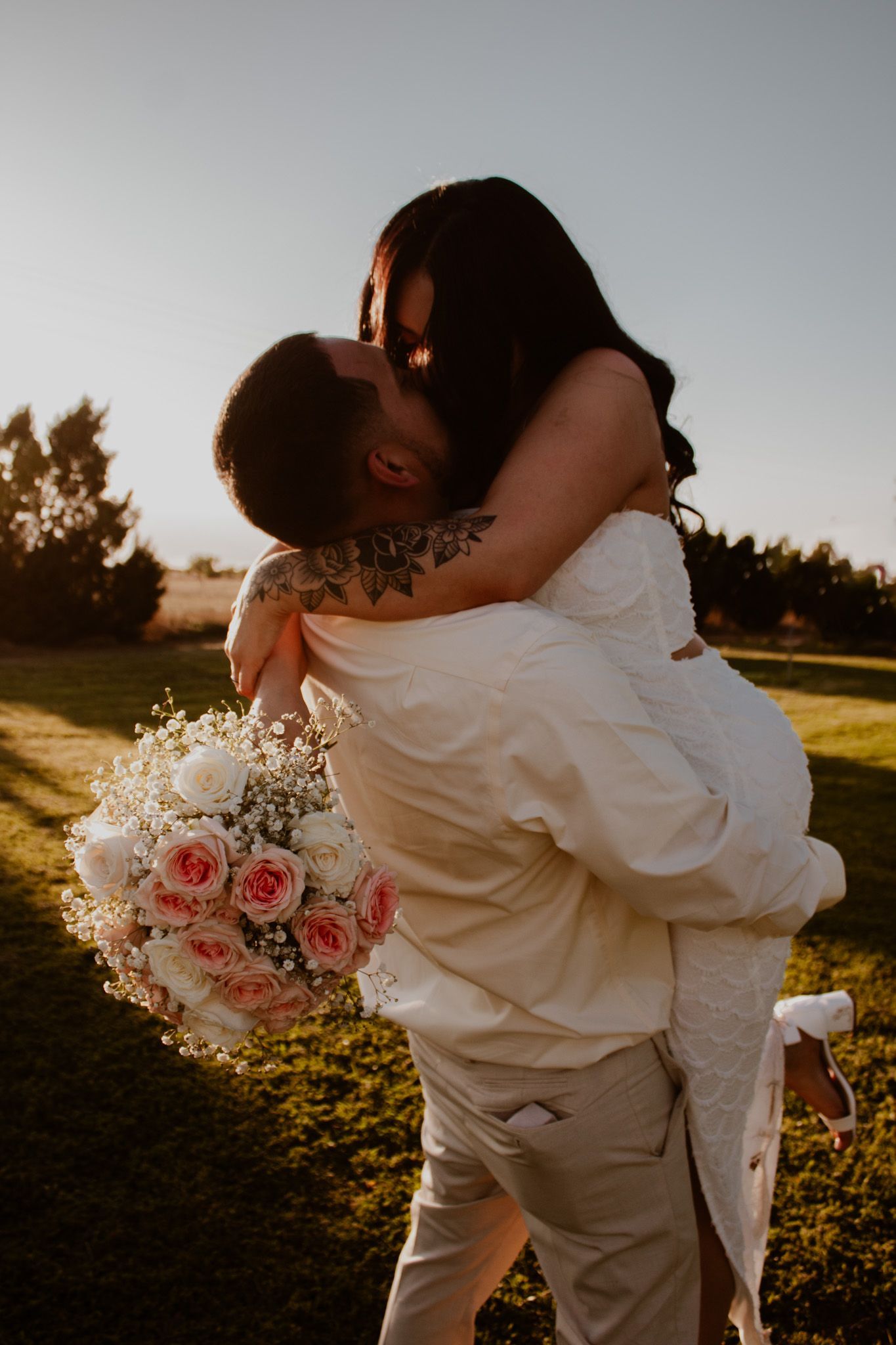 Groom lifts bride in white dress while kissing outdoors, she holds pink rose bouquet at golden hour wedding.
