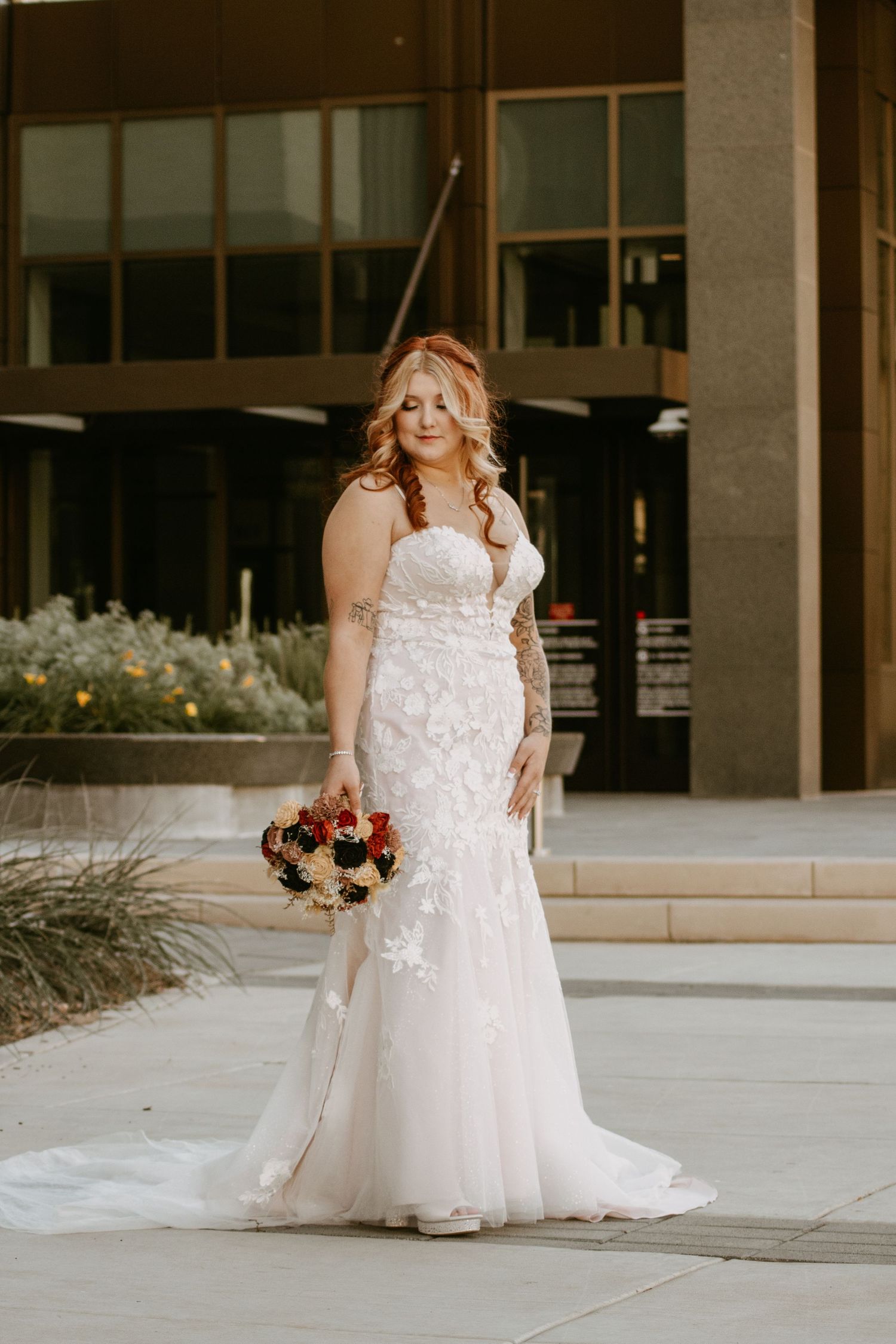 Bride in strapless lace mermaid wedding gown holding red and white bouquet standing outside modern building.