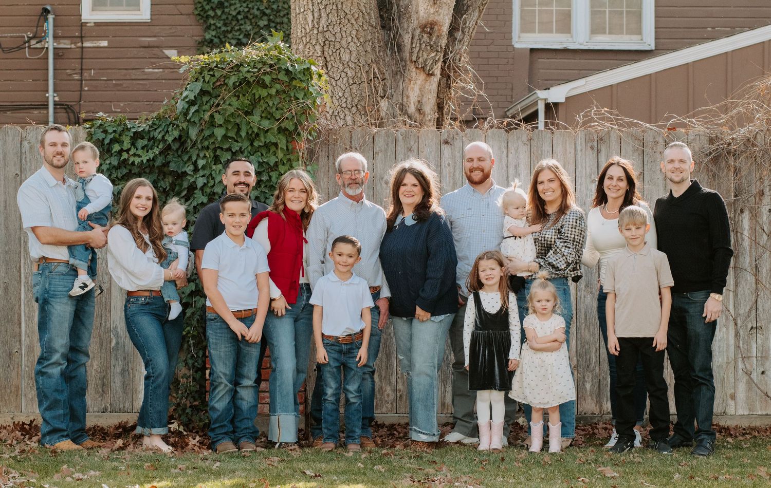 Large multigenerational family posing together outdoors in front of a wooden fence during a fall family portrait session.