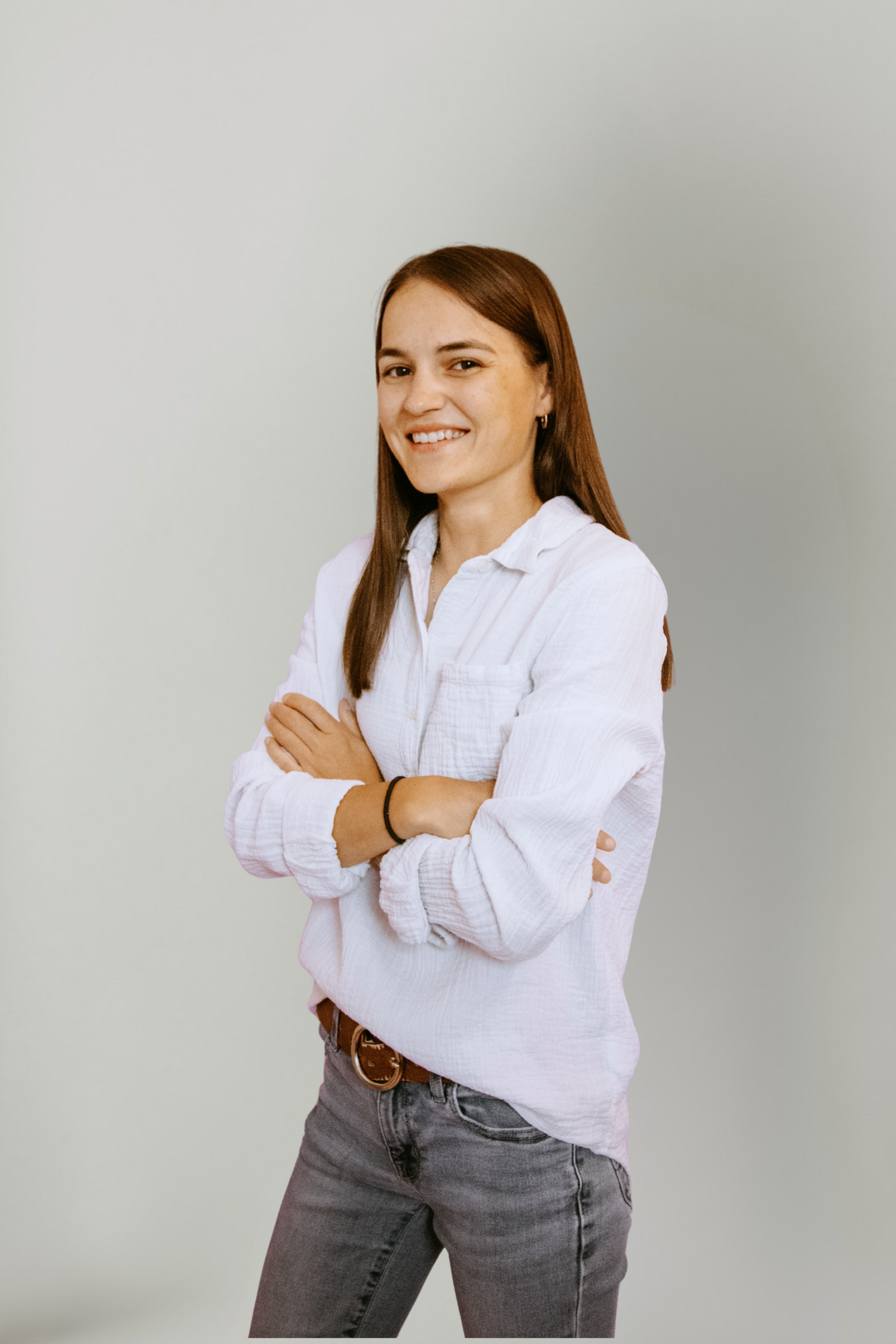 Smiling young woman in white shirt and jeans standing with arms crossed against a light gray background.