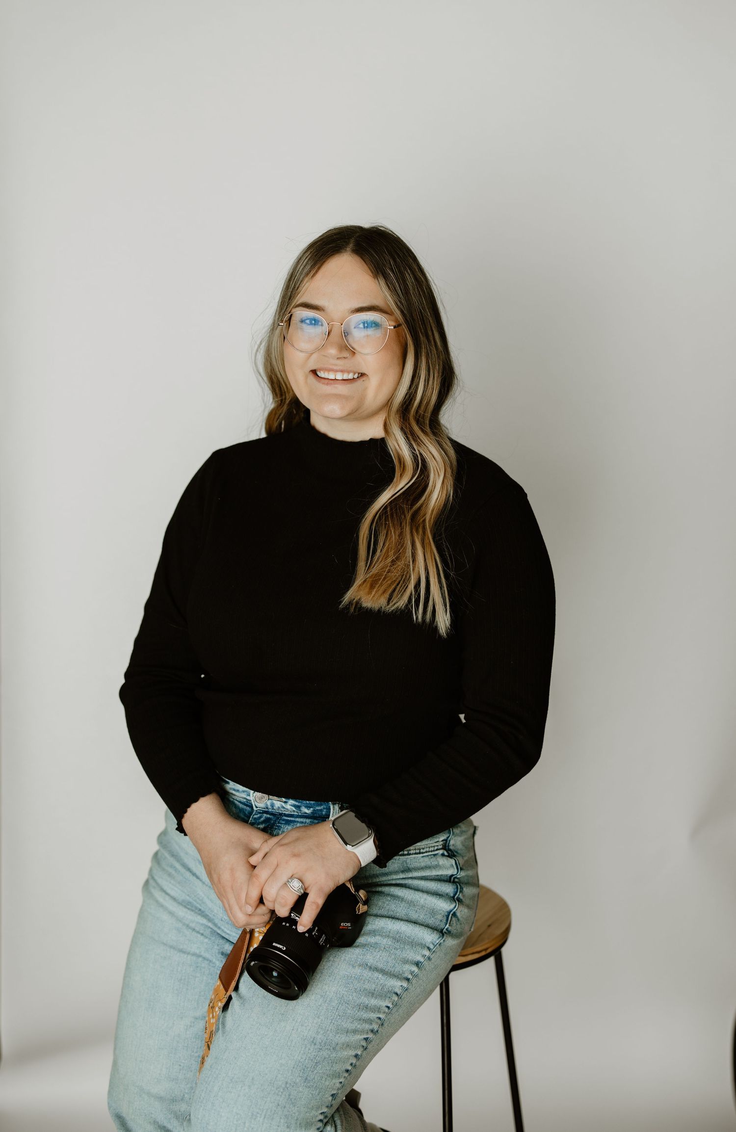 Smiling woman with glasses holding a camera, wearing black sweater and jeans, seated on stool against white background.