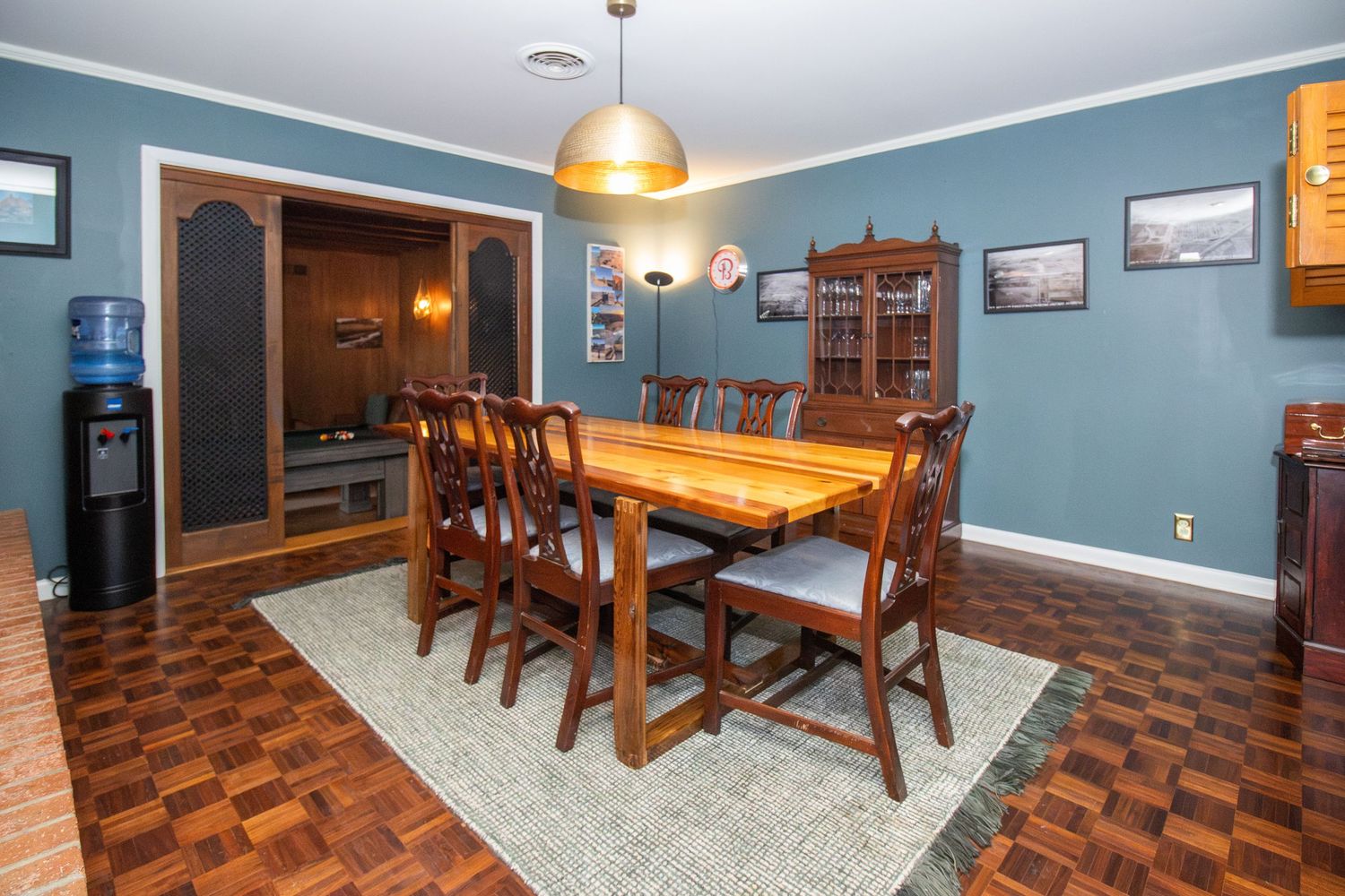 Elegant dining room with wooden table, chairs on herringbone parquet floor, blue walls, hutch, and water cooler.