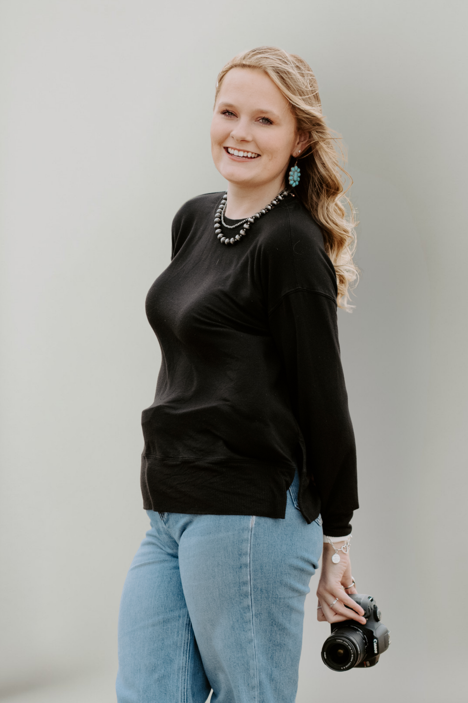 Smiling blonde woman in black top and jeans holding a camera, wearing turquoise earrings and necklace.