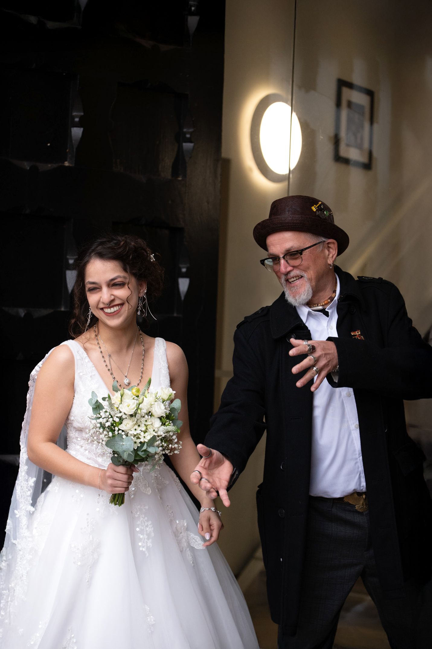 Smiling bride in white gown holding bouquet walks with older man in black coat and brown hat at wedding venue.