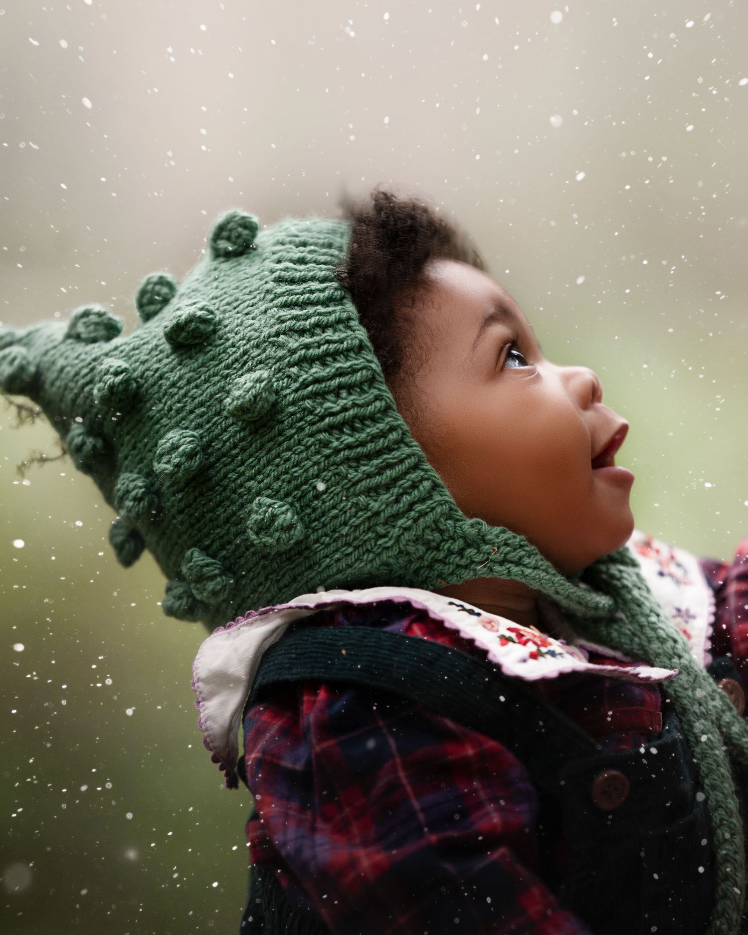 A child wearing a green knit hat with pom poms looks up with wonder as snow falls on a winter day.