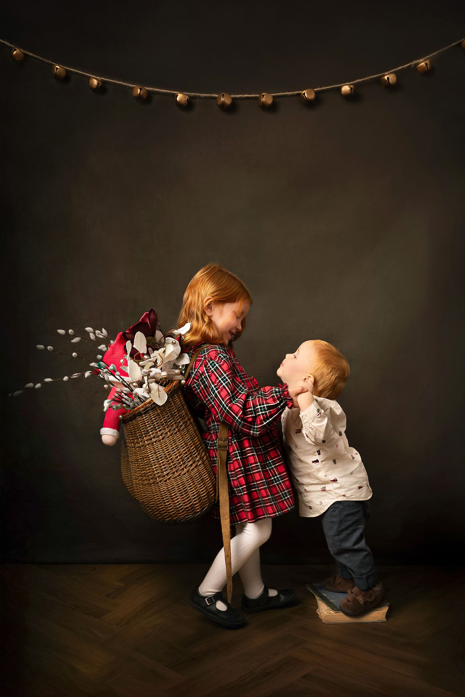 Two small children in holiday attire lean back-to-back while holding a wicker basket with decorative lights above.