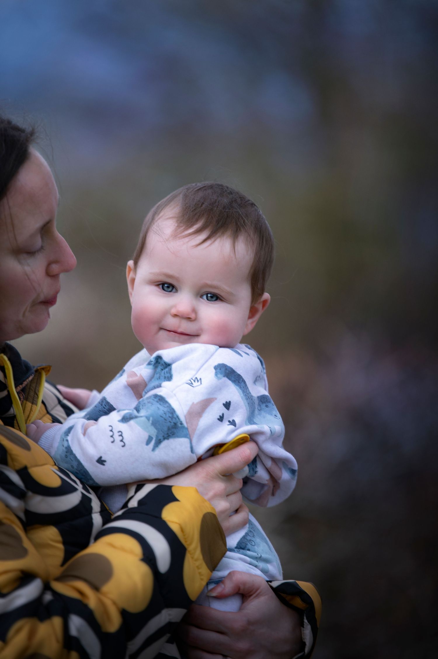 A baby wearing a white patterned sweater is being held lovingly outdoors during a winter day.