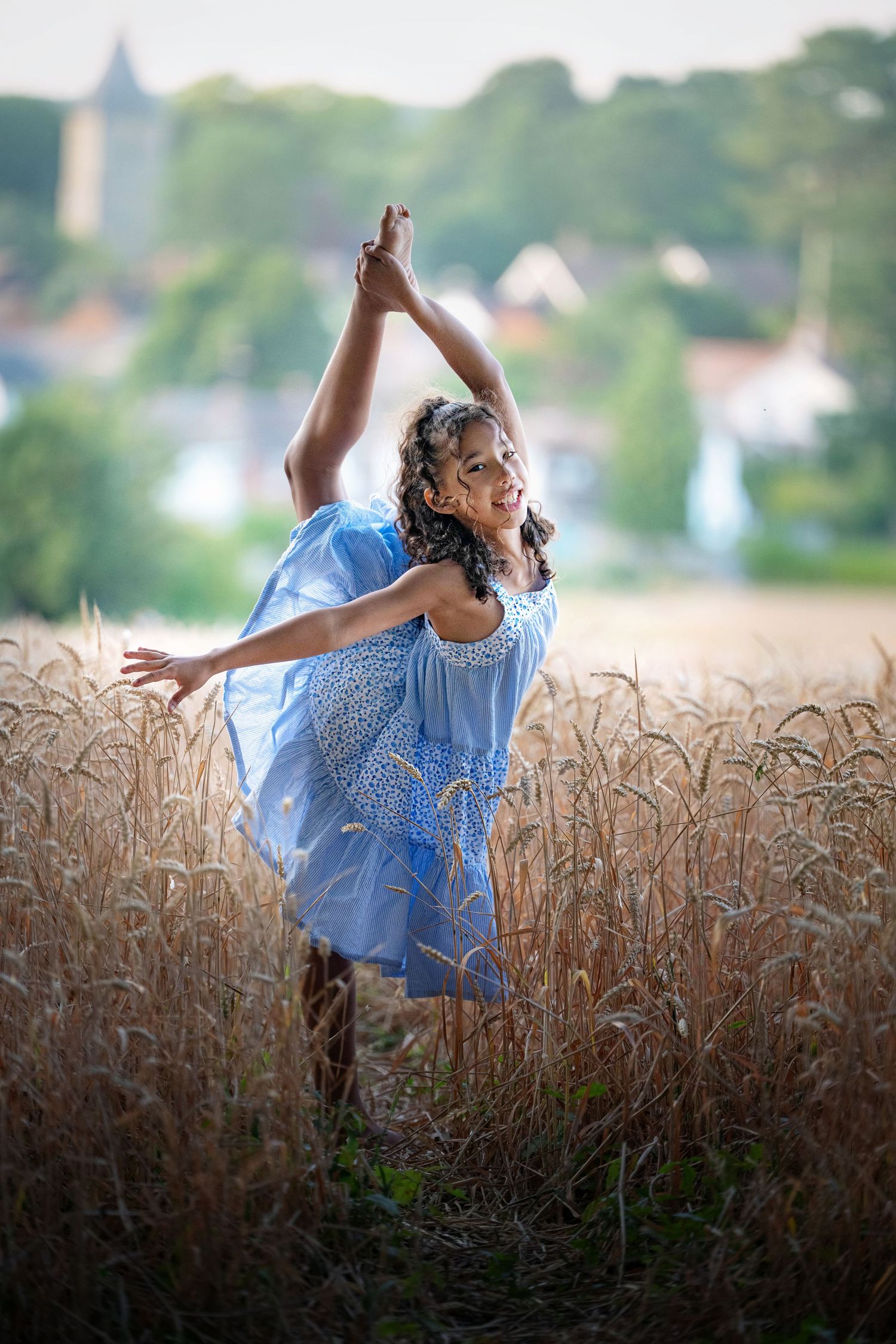 A woman in a blue dress performs a graceful dance pose in a golden wheat field with a village in the background.