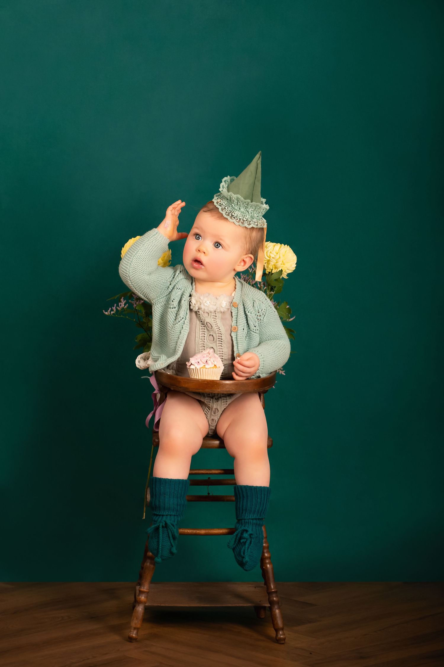 Cute baby in party hat sits on wooden chair with birthday cupcake against teal background.