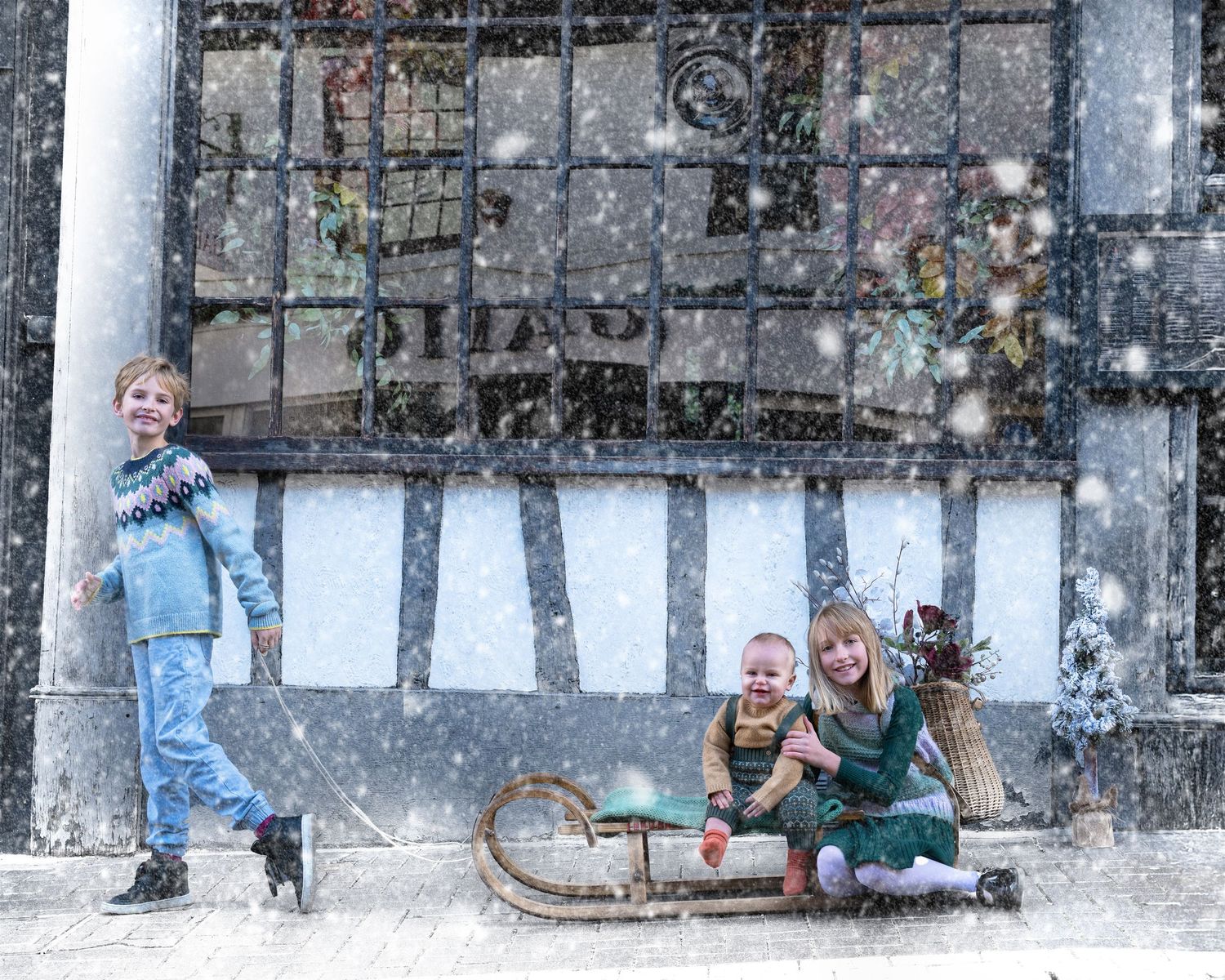Children enjoying winter fun on a wooden sled during a snowy day against a rustic wall backdrop.