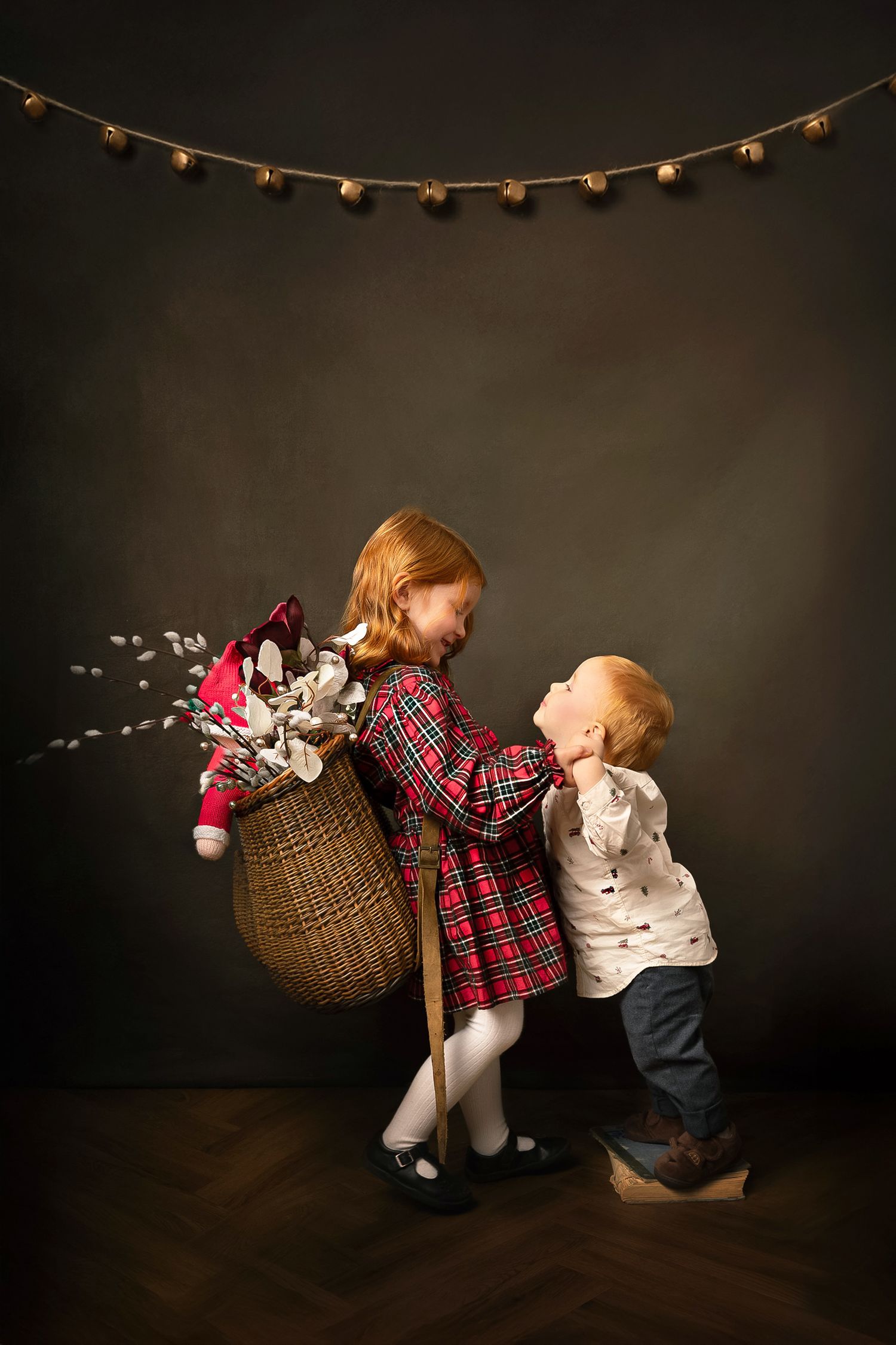 Two children in plaid and white outfits lean and play under string lights in a dark studio setting.