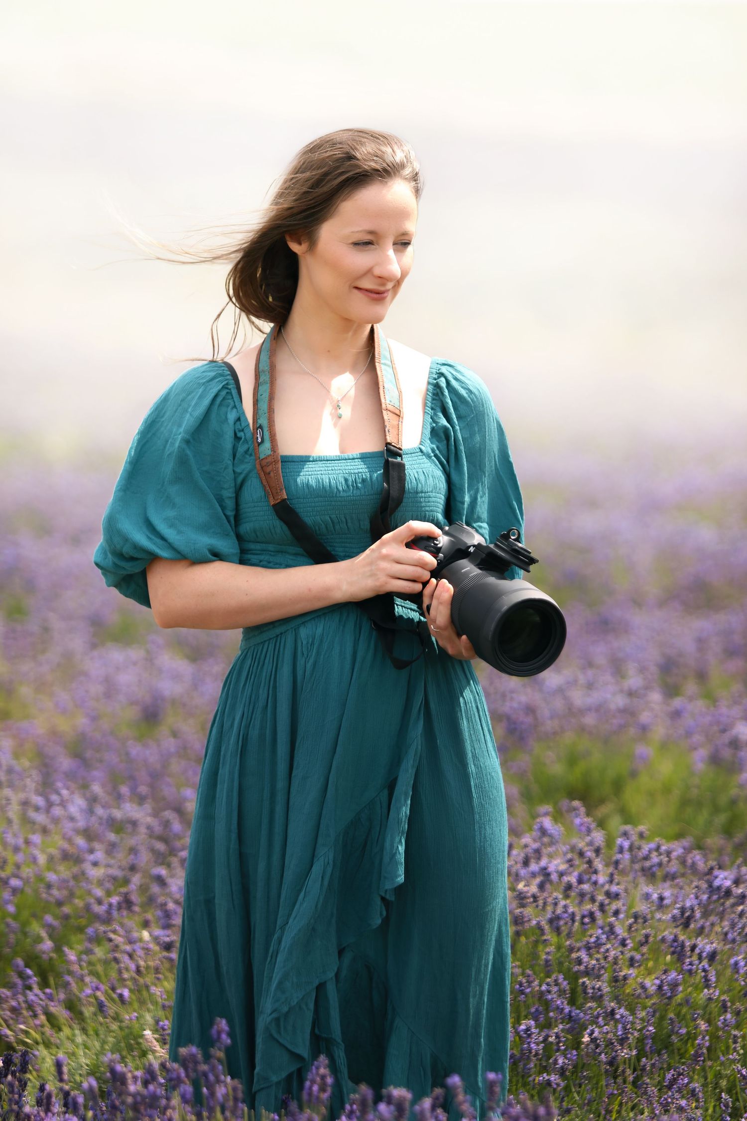 Woman in teal dress holding DSLR camera smiling in purple lavender field on a misty day.