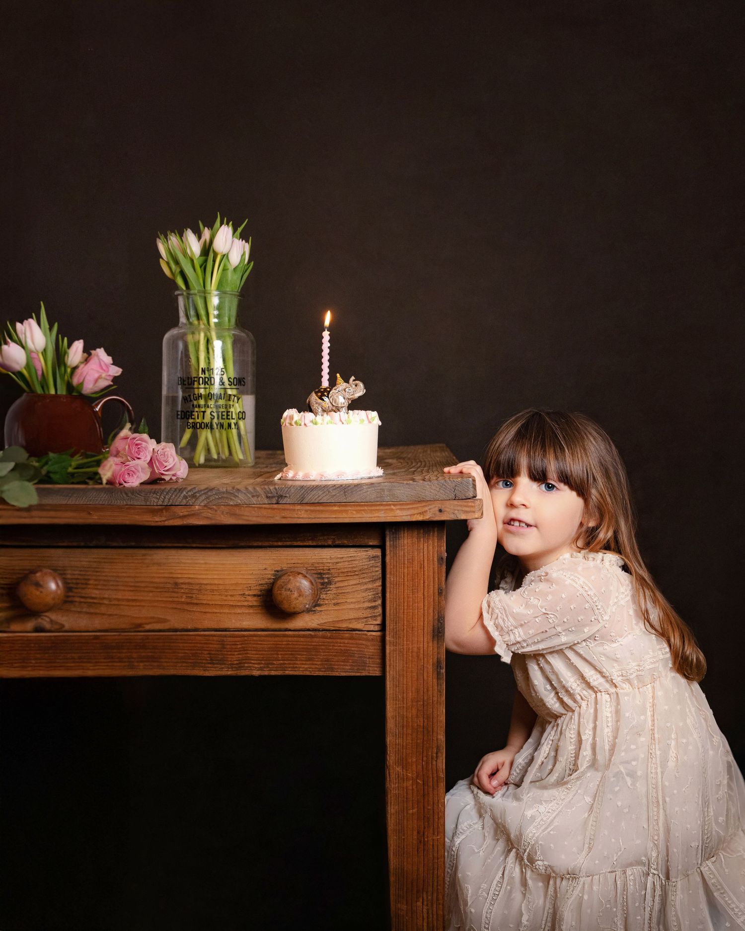Young girl in white lace dress stands beside wooden table with birthday cake and pink tulips against dark background.
