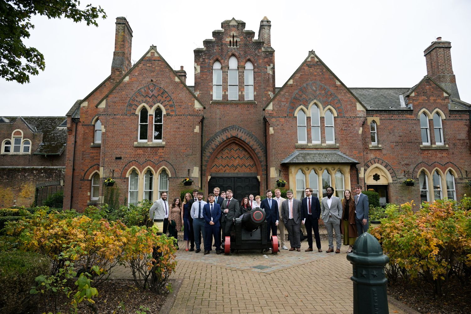 Group of professionals posing outside a historic red brick Gothic building with arched doorway and manicured gardens.