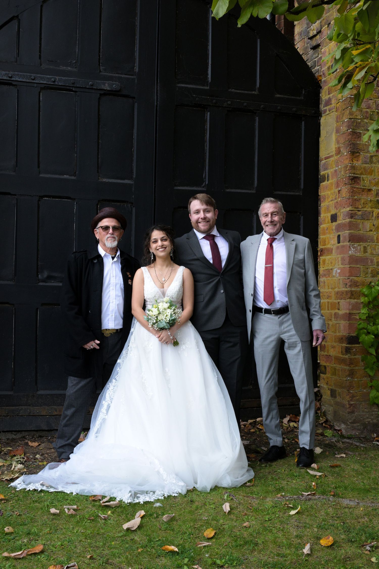 Bride in white gown holds bouquet posing with three men in suits before black doors at outdoor wedding.