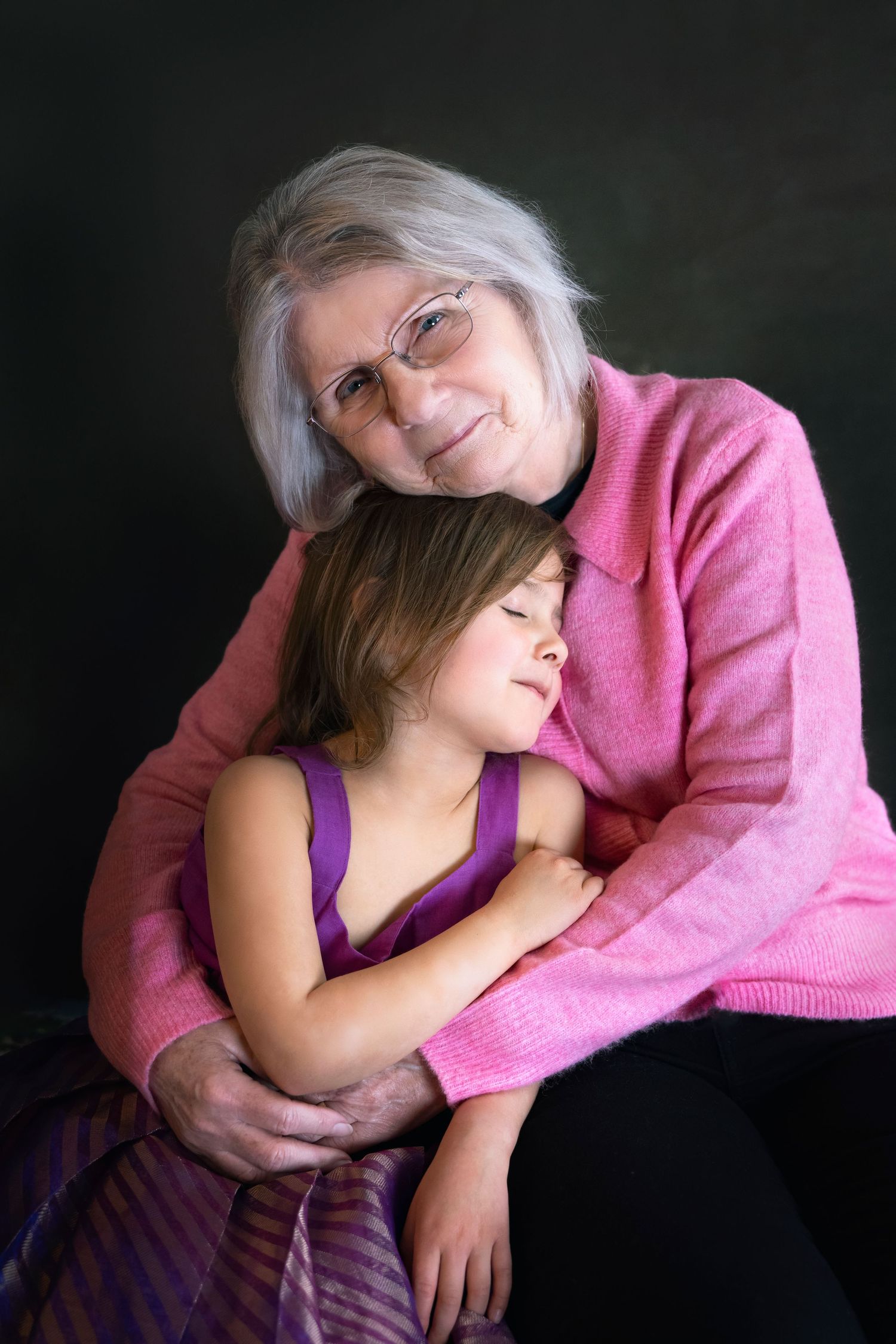 Elderly grandmother with gray hair and glasses tenderly embracing a young girl in a studio portrait.