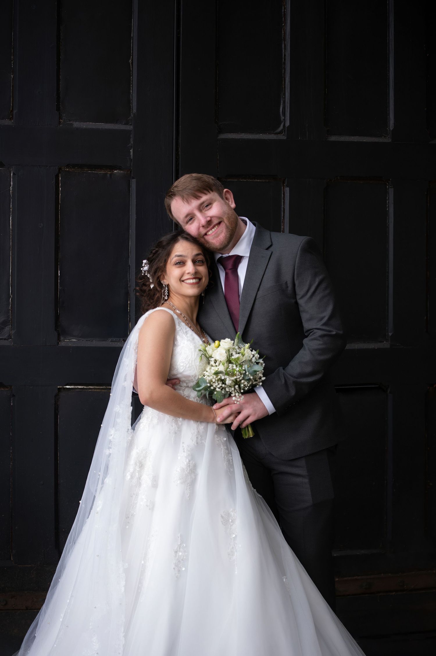 Smiling bride in white gown holding bouquet with groom in gray suit posing together against dark wood backdrop.