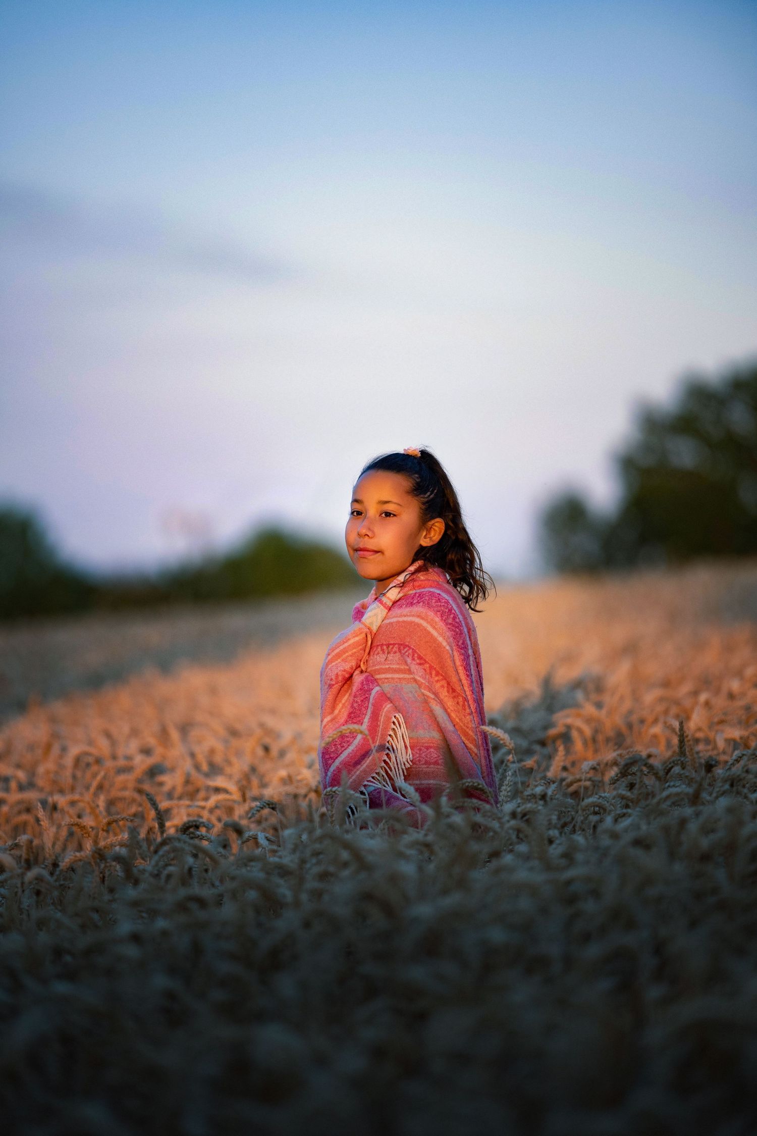 Young girl wrapped in colorful shawl standing in golden wheat field at dusk with soft blue sky background.